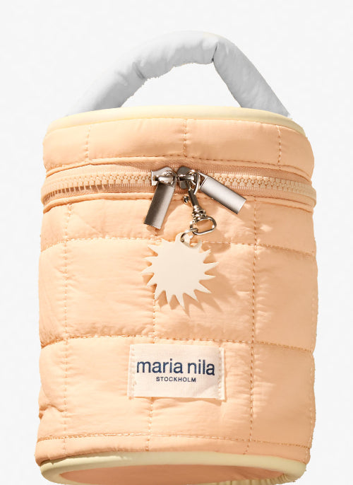 A light peach quilted puffer bag by Maria Nila, featuring a top zipper with silver hardware, a white sun-shaped charm, and a white fabric label with the brand name.