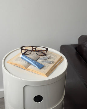 Round white side table with an open book, glasses, and a blue object on top, next to a dark leather sofa.